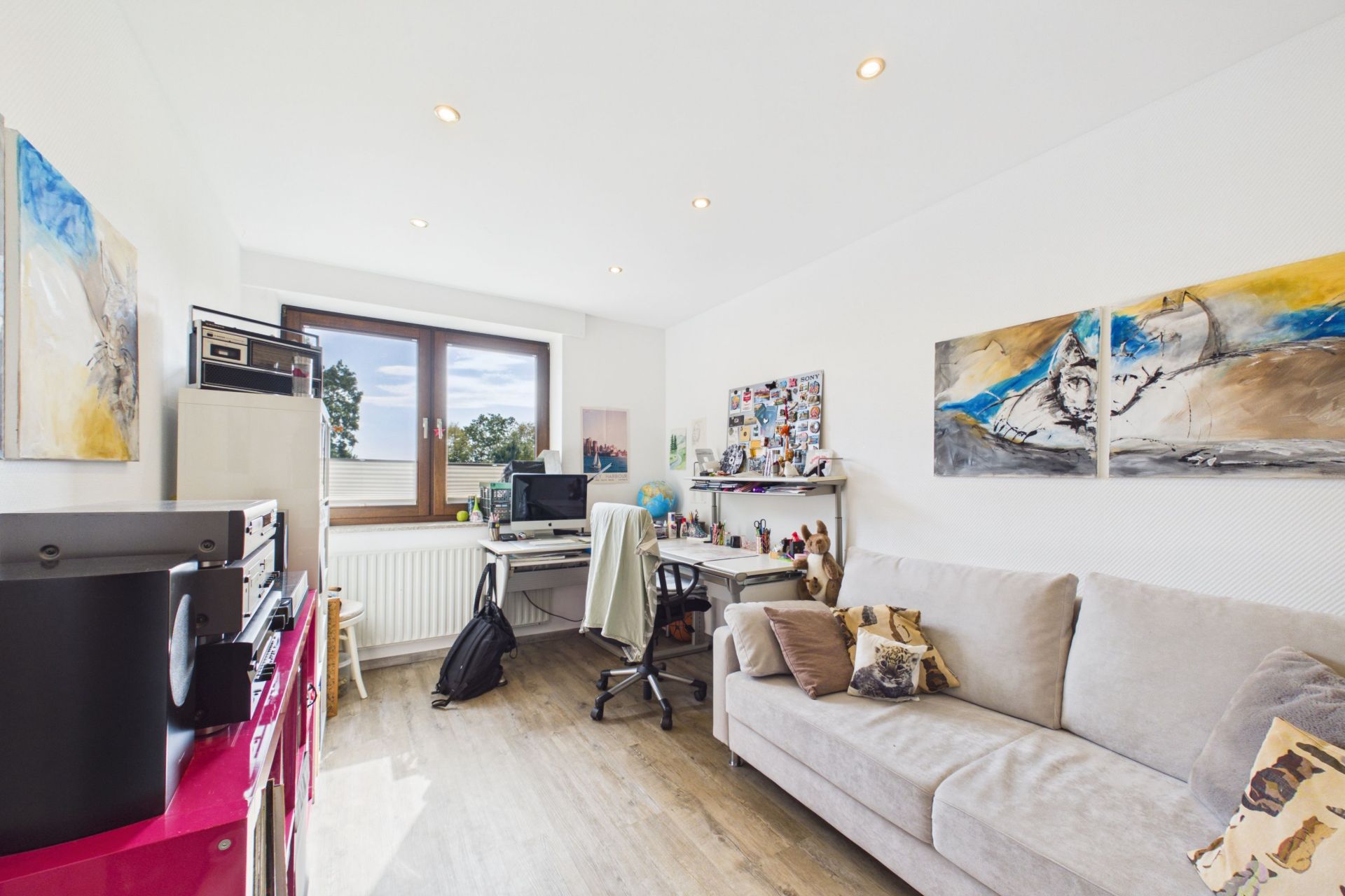 Helles Arbeitszimmer mit Sofa, Schreibtisch, Computer und großen Fenstern.