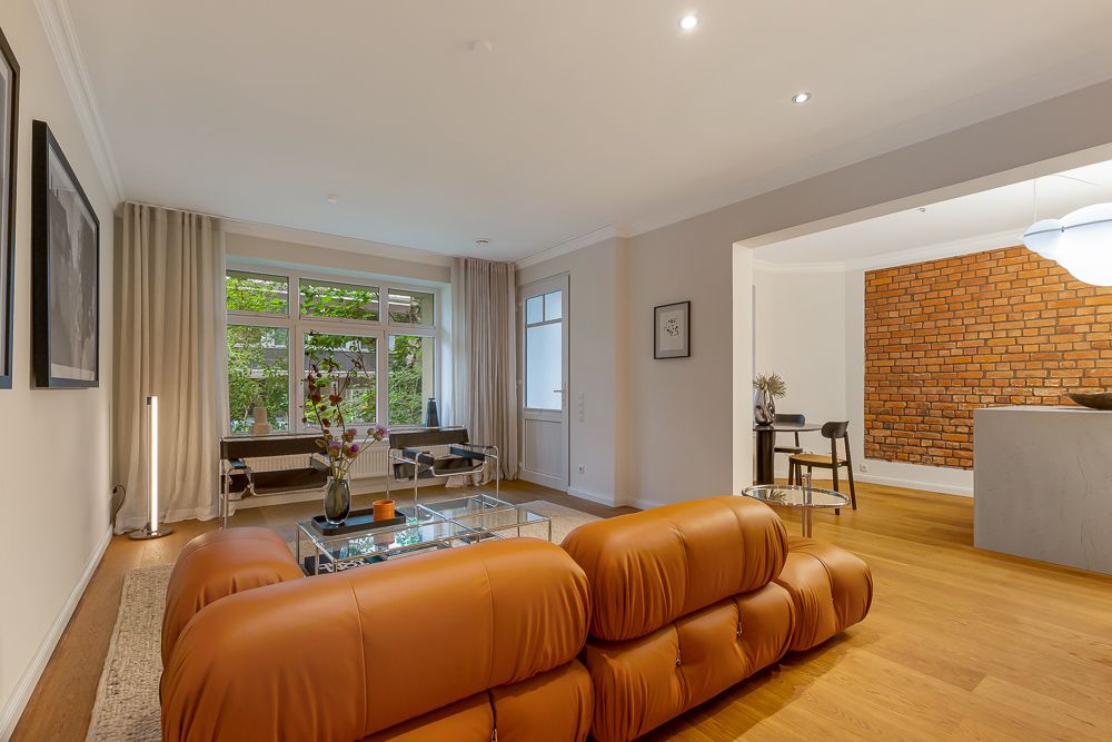 Modernes Wohnzimmer mit braunem Sofa, Glastisch, und großer Fensterfront, Blick auf Essbereich mit Backsteinwand.