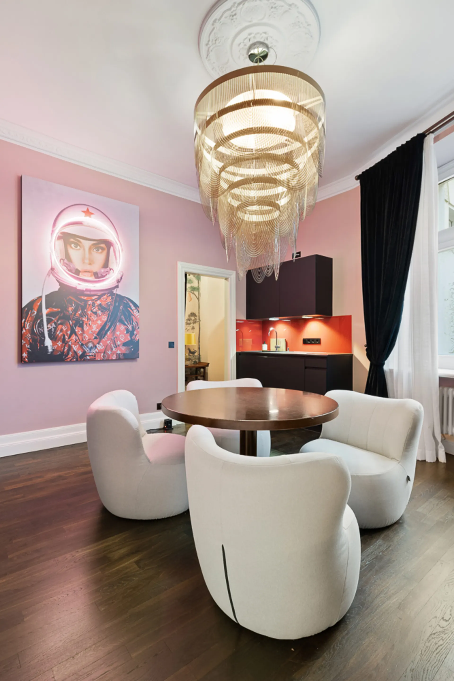 Wohnzimmer mit rundem Holztisch, vier weißen Sesseln, großem Kronleuchter und Wandbild.