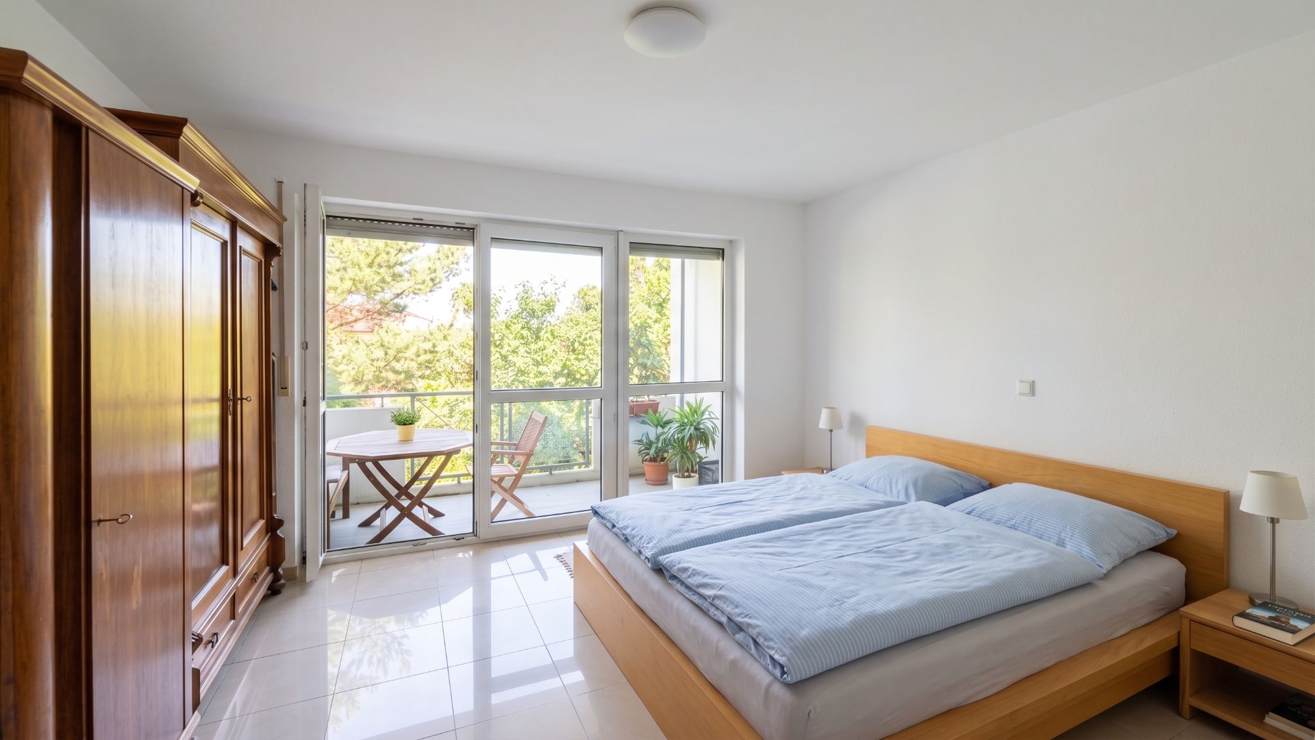 Helles Schlafzimmer mit Doppelbett, Holzschrank und Zugang zu einem Balkon mit Sitzgruppe.