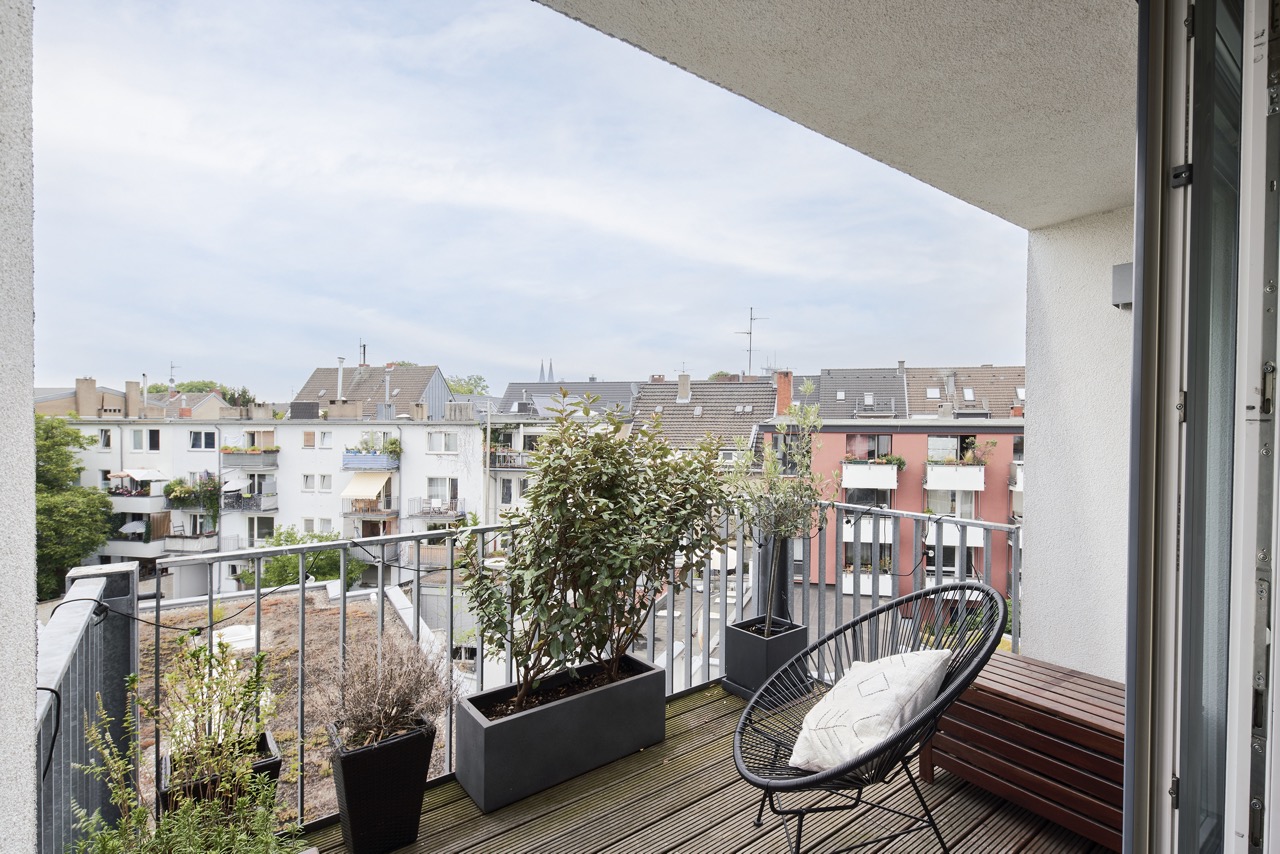 Balkon mit Pflanzen, Stuhl mit Kissen und Bank, Blick auf andere Wohnhäuser.