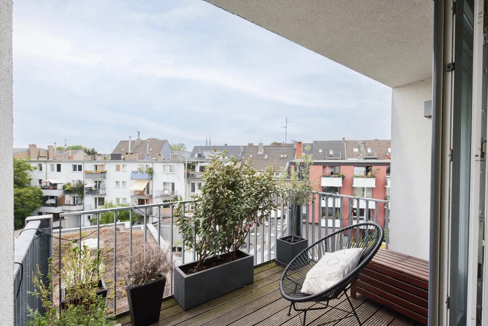 Balkon mit Pflanzen, Stuhl mit Kissen und Bank, Blick auf andere Wohnhäuser.