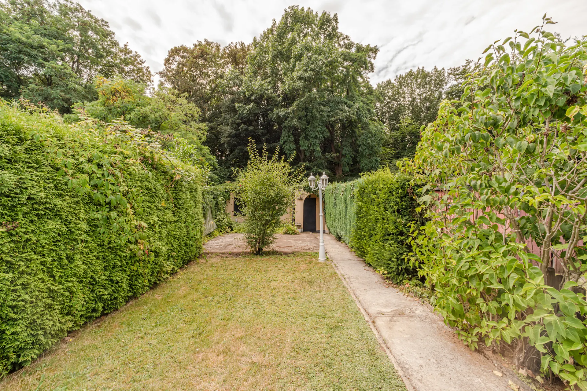 Garten mit Rasenfläche, Gehweg, Hecken und einer kleinen Hütte im Hintergrund.