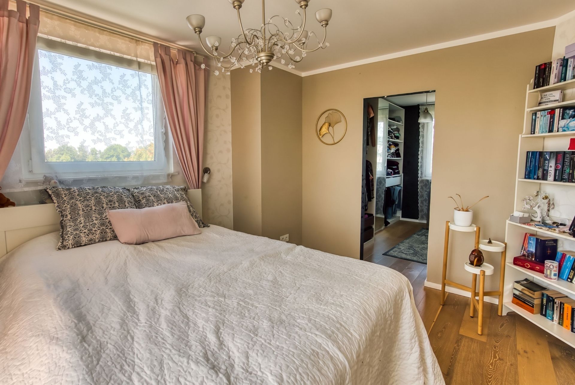 Schlafzimmer mit Doppelbett, Kronleuchter, Bücherregal und Fenster mit rosa Vorhängen.