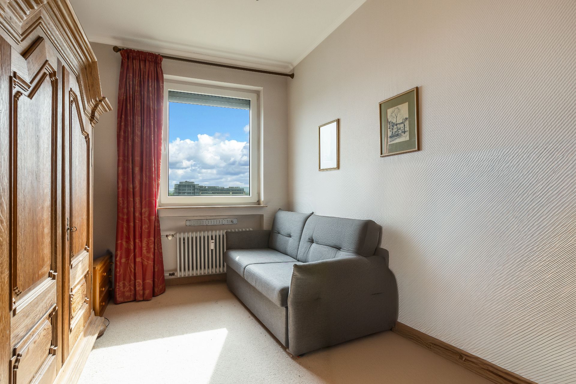 Kleines Zimmer mit grauem Sofa, Holzschrank, Fenster und zwei Bildern an der Wand.