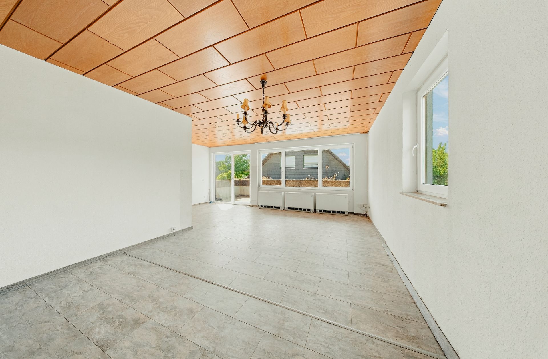 Heller, leerer Raum mit Fliesenboden, Holzelementdecke, Kronleuchter und großen Fenstern.