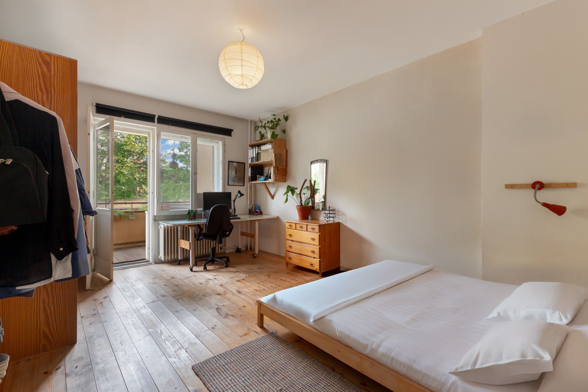 Helles Schlafzimmer mit Bett, Schreibtisch, Kleiderschrank und offenem Fenster zum Balkon.