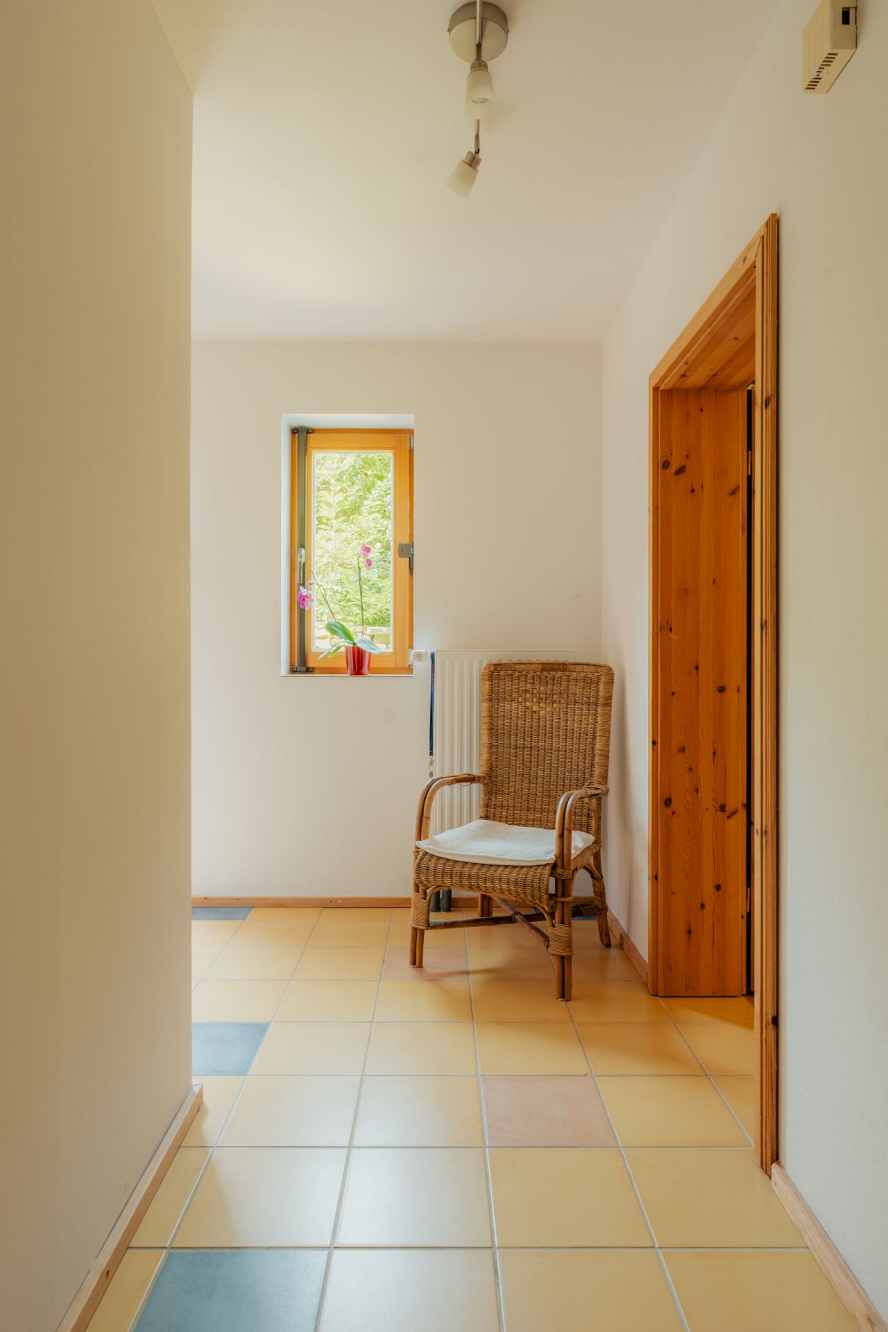 Flur mit gefliestem Boden, Korbstuhl, Fenster mit Blume und Holztürrahmen.