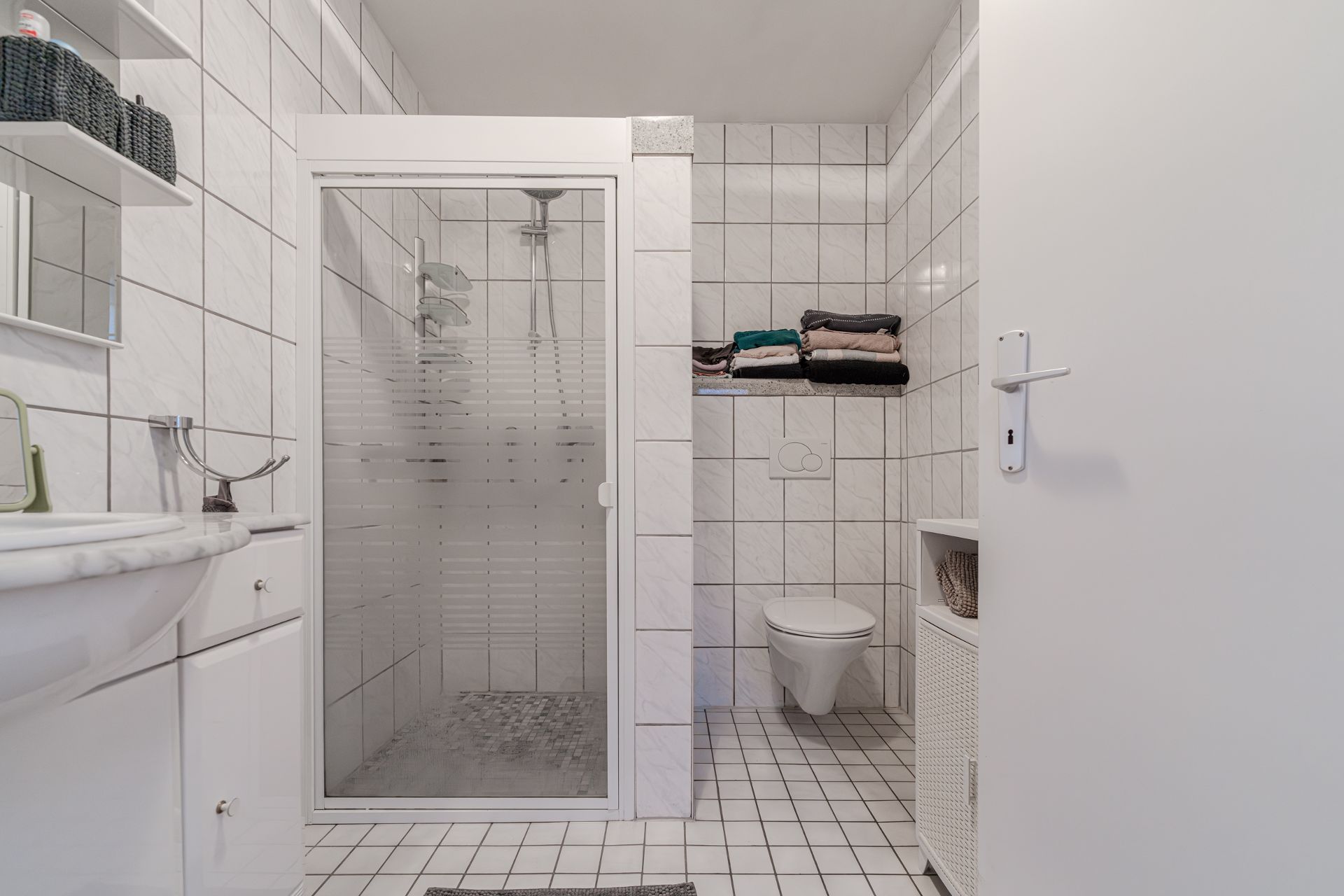 Modernes, gefliestes Badezimmer mit Dusche, Toilette, Waschbecken und Regalfächern mit Handtüchern.