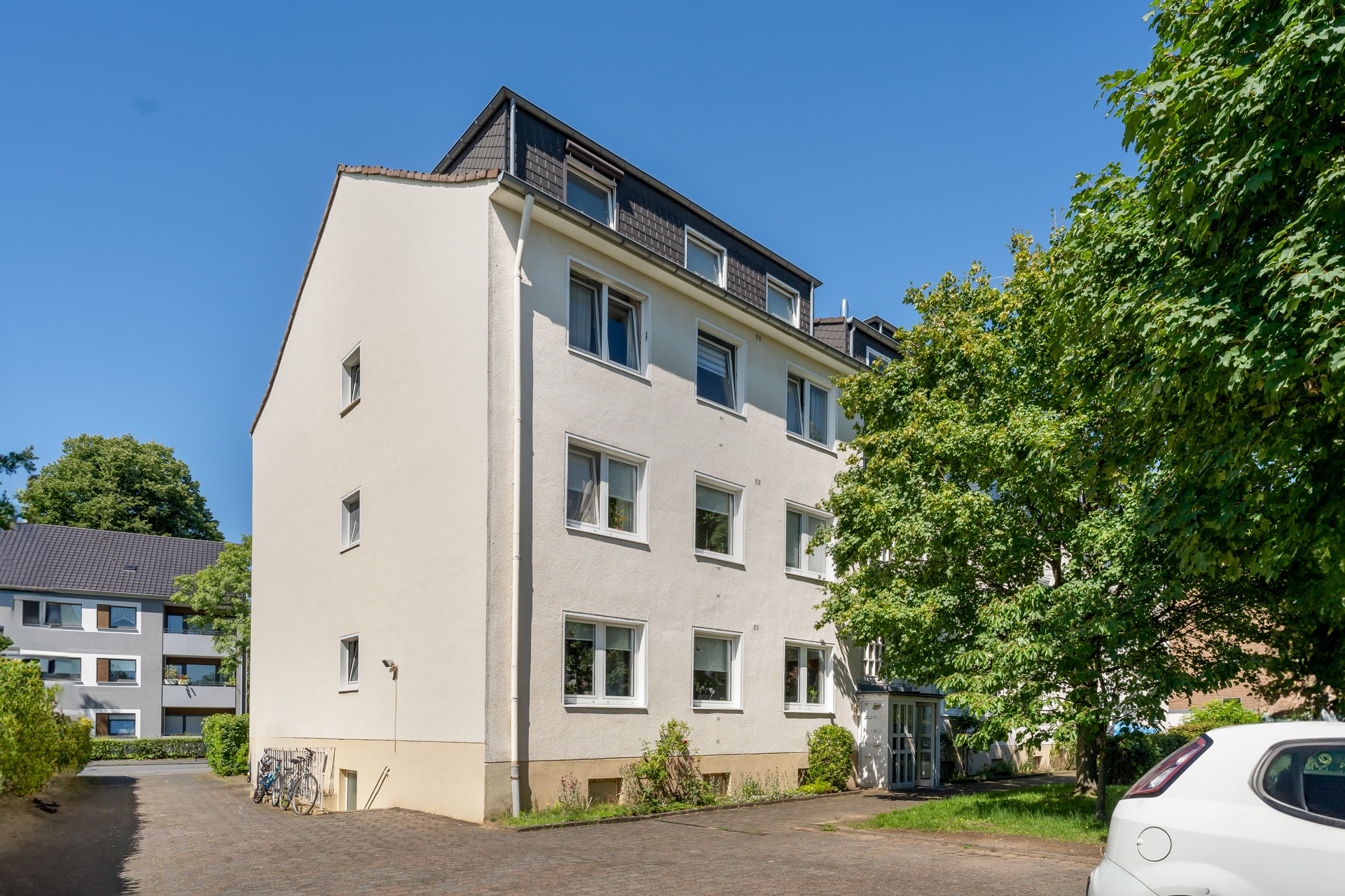 Mehrfamilienhaus mit weißer Fassade, mehreren Fenstern und Bäumen davor.