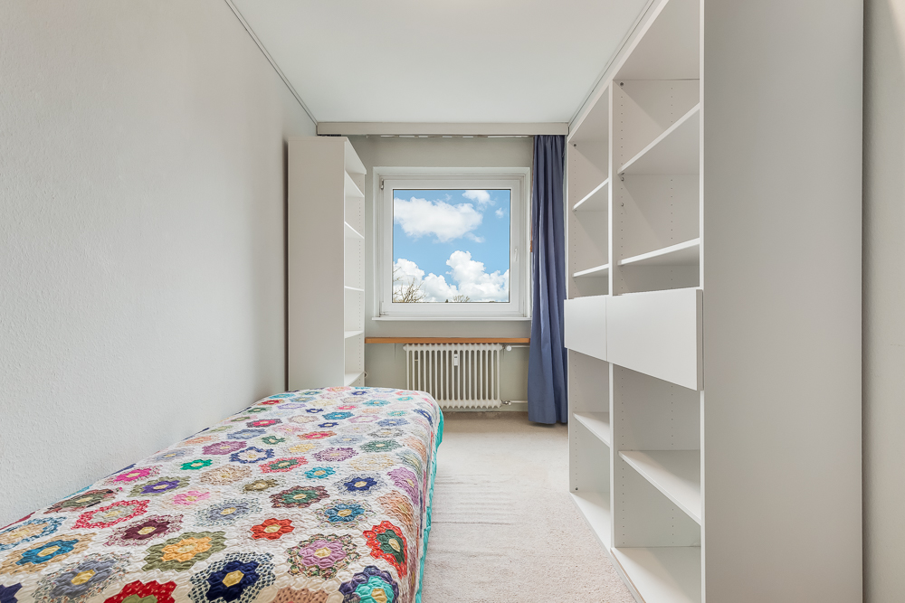 Kleines Schlafzimmer mit Einzelbett, bunter Decke, weißen Regalen und Fenster mit blauen Vorhängen.