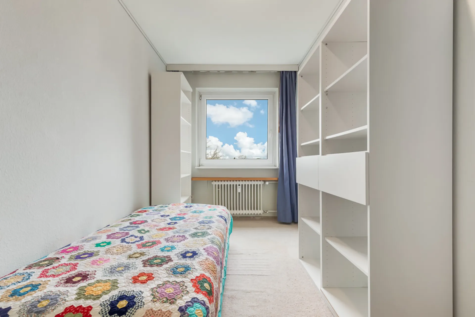 Kleines Schlafzimmer mit Einzelbett, bunter Decke, weißen Regalen und Fenster mit blauen Vorhängen.