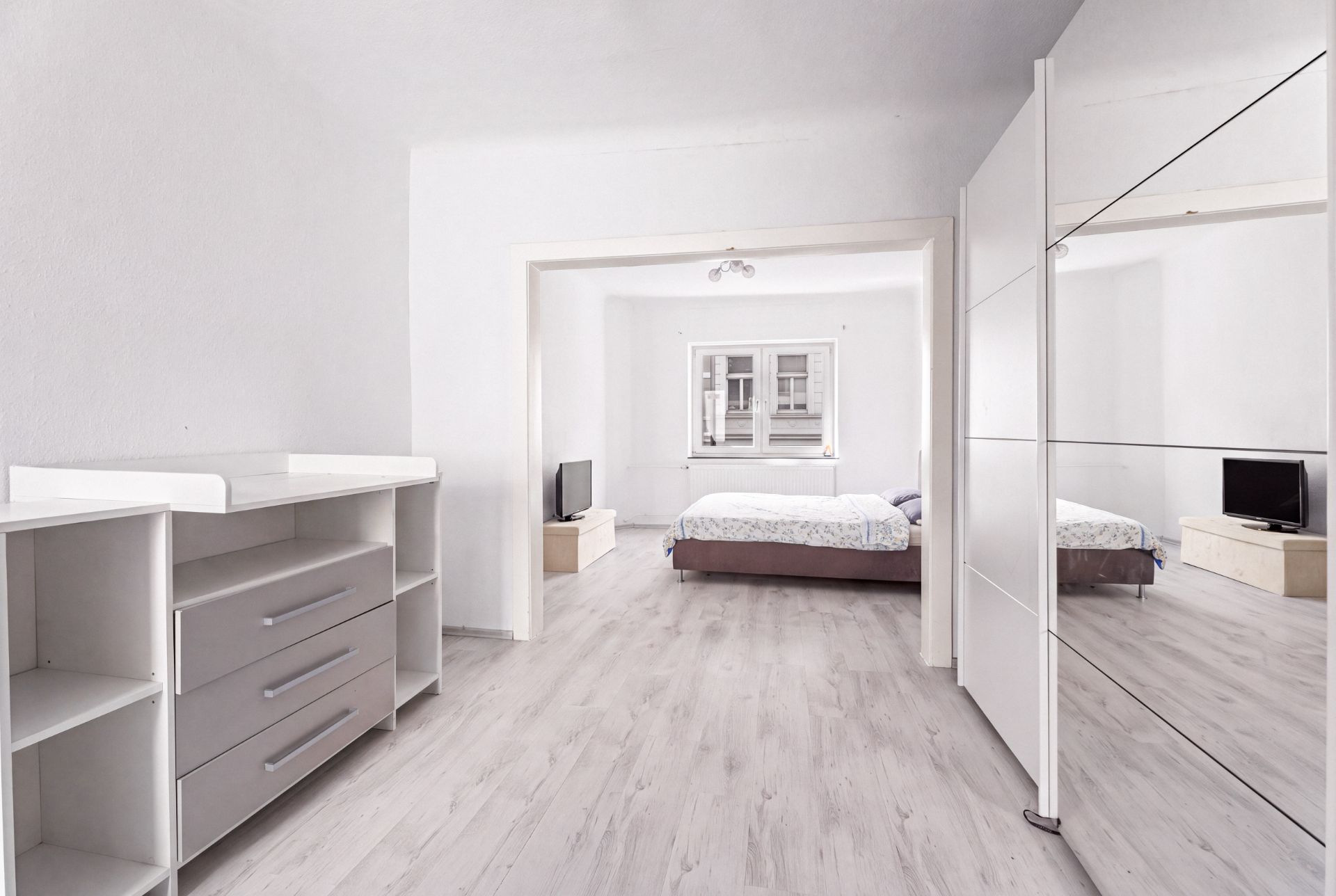 Helles Schlafzimmer mit Doppelbett, Kommode, Spiegel und Fernseher, Blick auf ein Fenster.