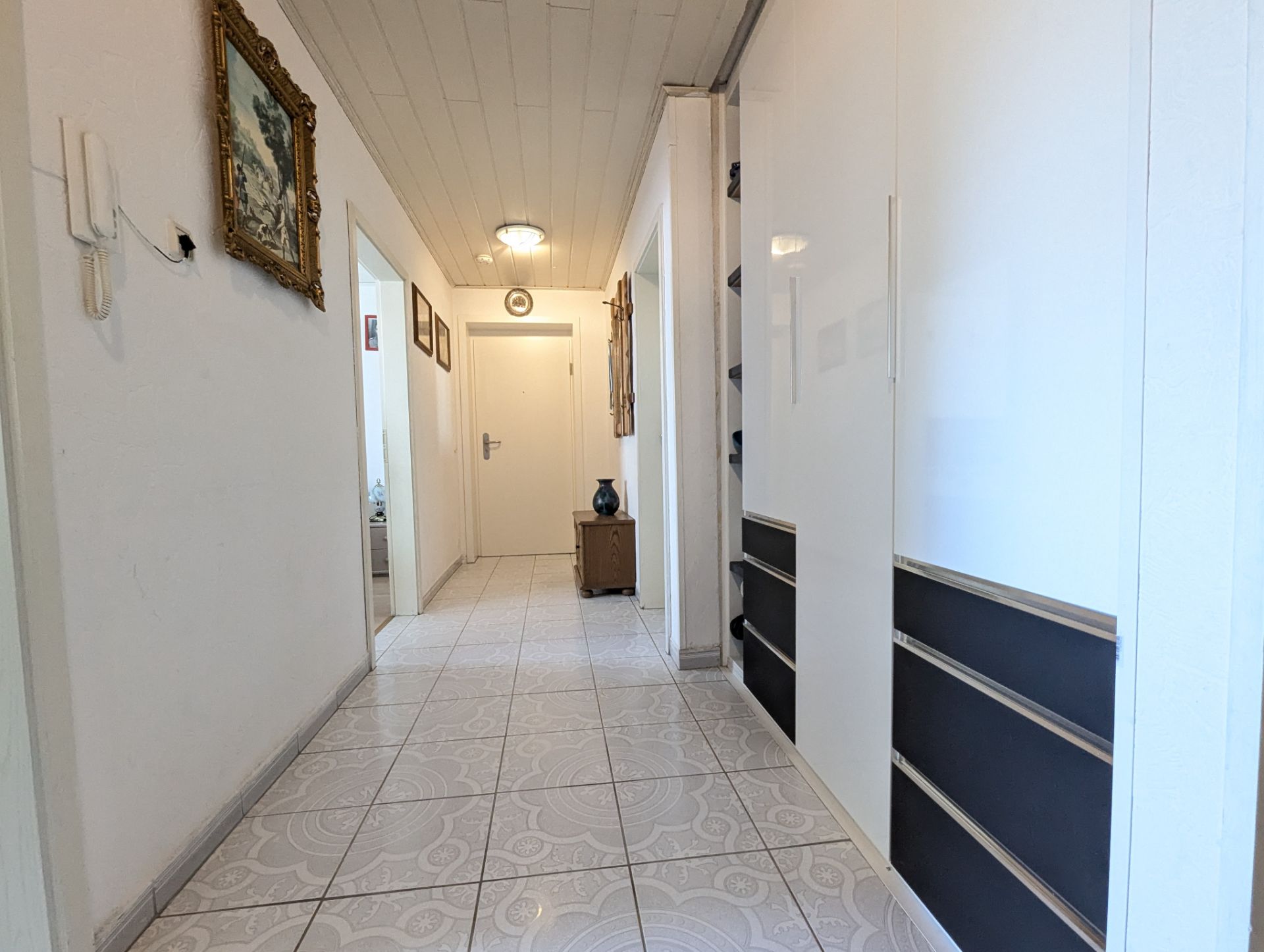 Heller Flur mit Fliesenboden, weißen Wänden, Bildern und Schrank.