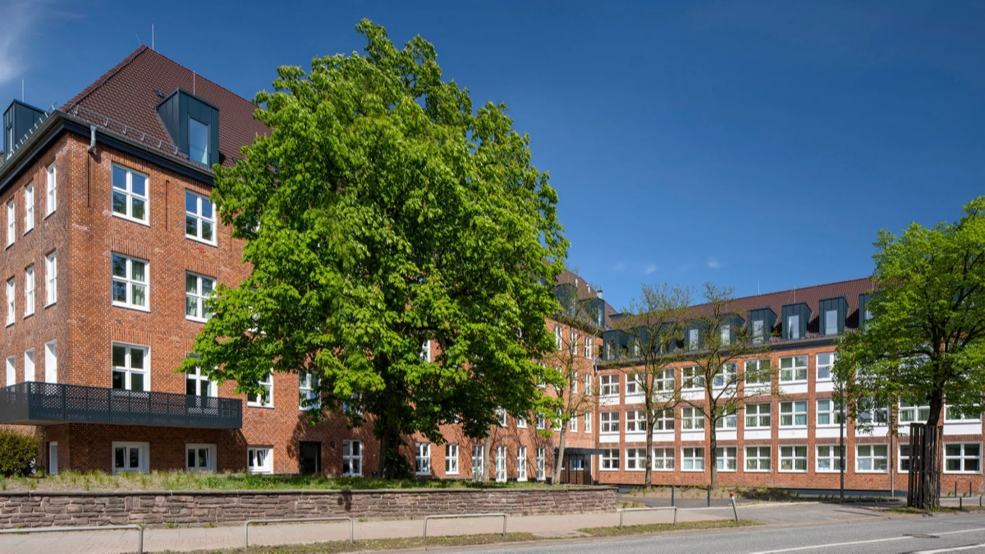 Großes, vierstöckiges Wohngebäude aus rotem Backstein mit vielen Fenstern und Bäumen davor.