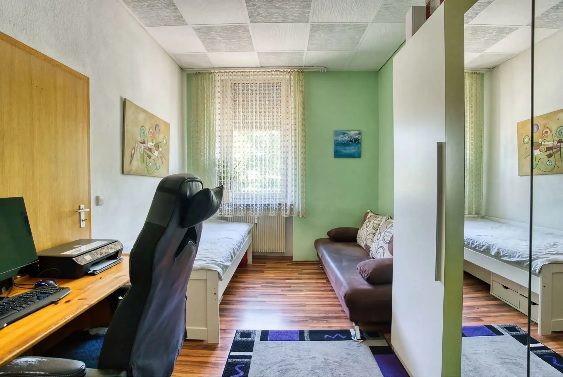 Zimmer mit zwei Betten, Schreibtisch mit Computer, Sofa, Fenster mit Gardine und Holzfußboden.