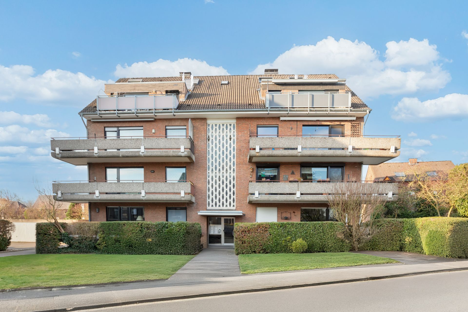 Mehrfamilienhaus mit Balkonen, roter Klinkerfassade und gepflegtem Vorgarten bei Tageslicht.
