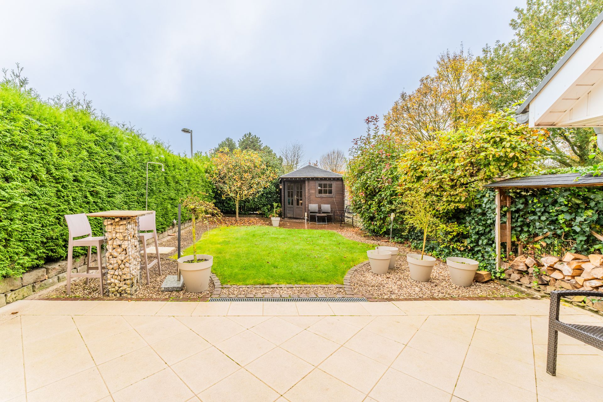 Gepflegter Garten mit Terrasse, Rasen, Sitzbereich und kleinem Holzhaus im Hintergrund.