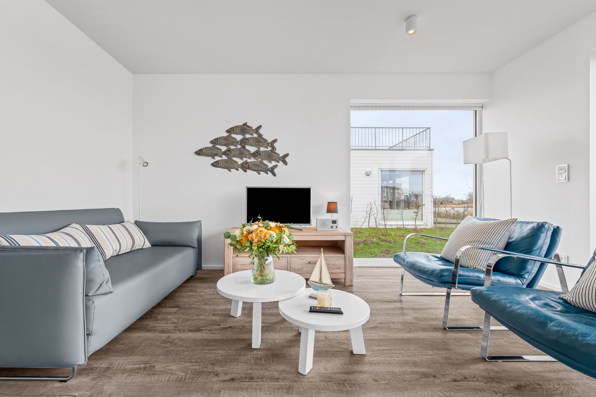 Modernes Wohnzimmer mit Sofa, zwei blauen Sesseln, Couchtischen, Fernseher und Glasvase mit Blumen.