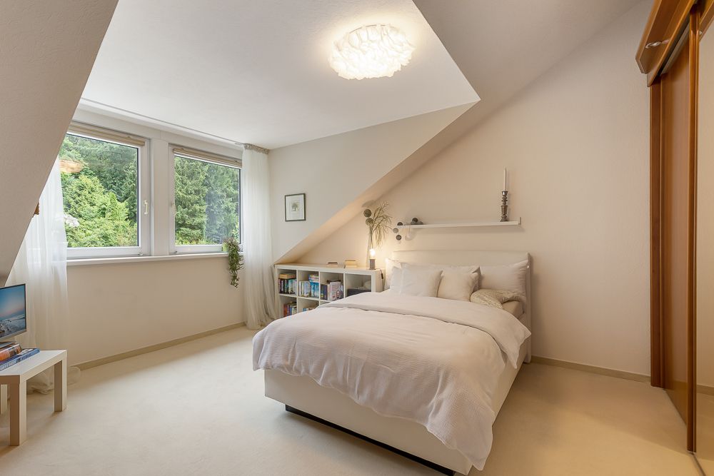 Helles Schlafzimmer mit Doppelbett, großem Fenster, Bücherregal und schräger Decke.