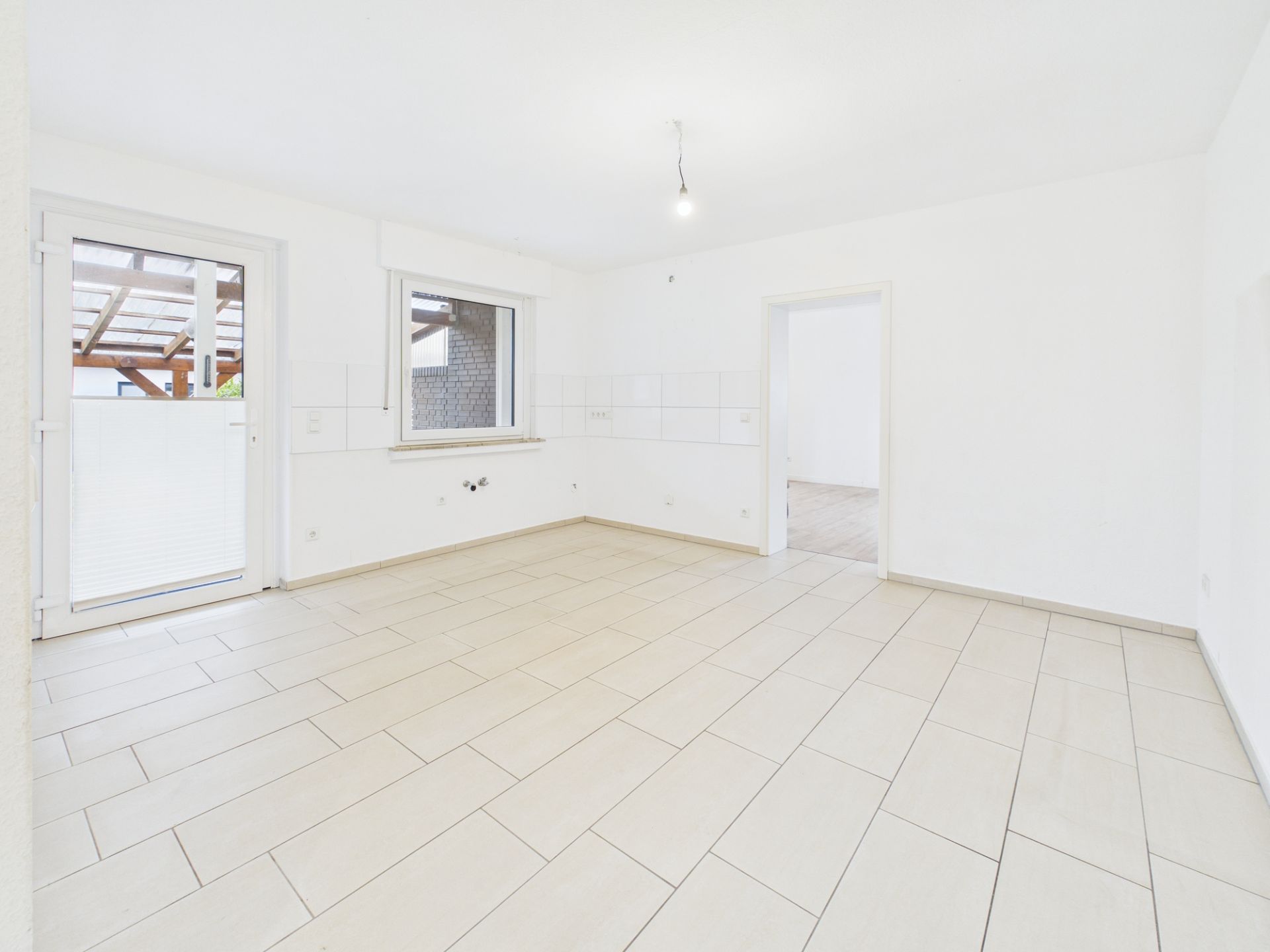 Leerer, heller Raum mit Fliesenboden, Fenster, Tür zum Balkon und Eingang zu einem weiteren Zimmer.
