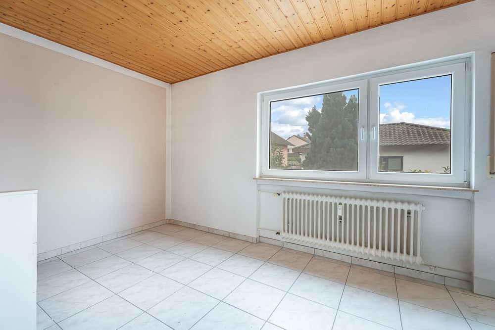 Leerer Raum mit weißem Fliesenboden, Holzdecke, großem Fenster und Heizkörper unter dem Fenster.
