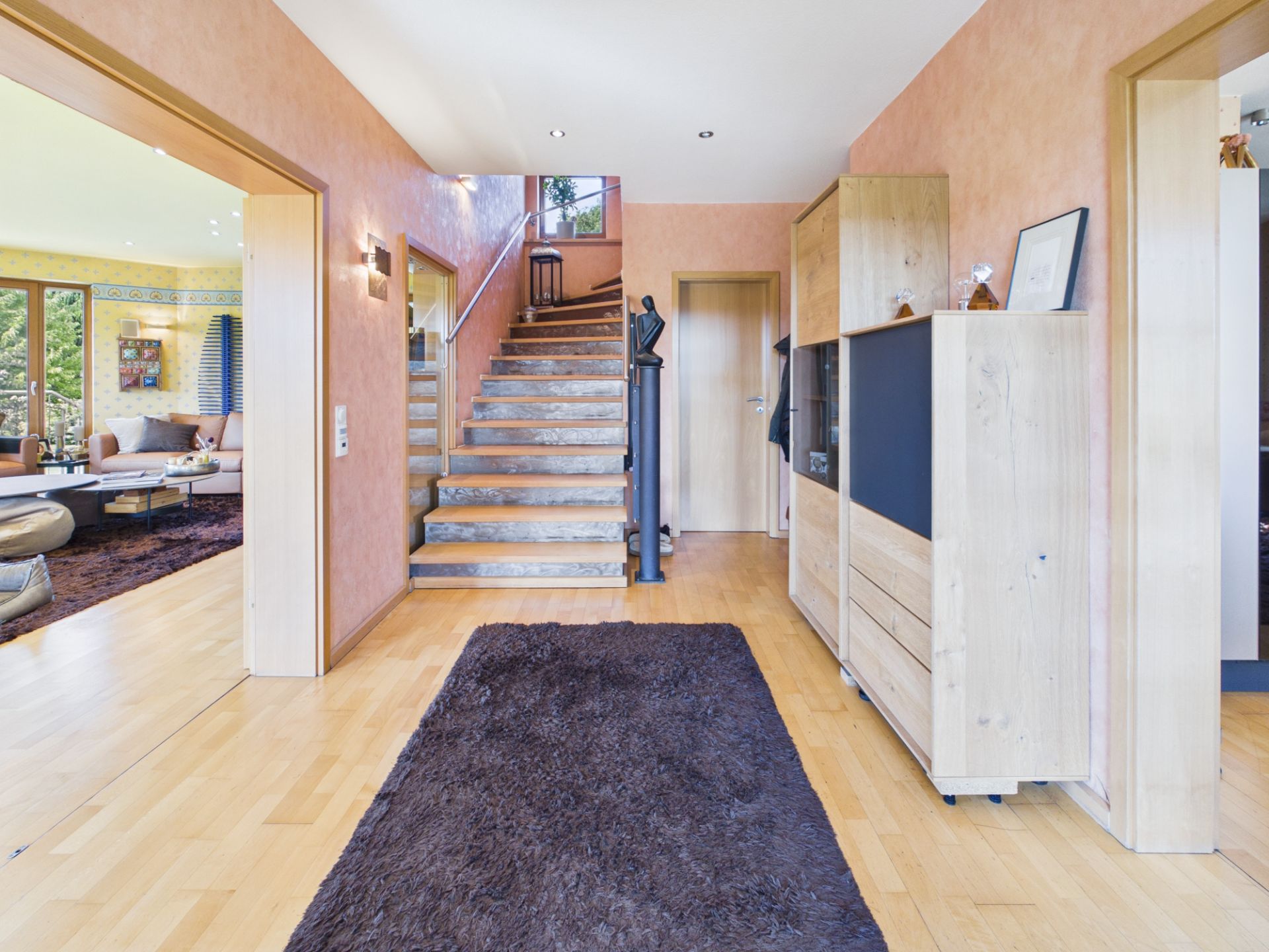 Heller Flur mit Holztreppe, Teppich und Holzschrank. Links ein Wohnzimmer mit großen Fenstern.