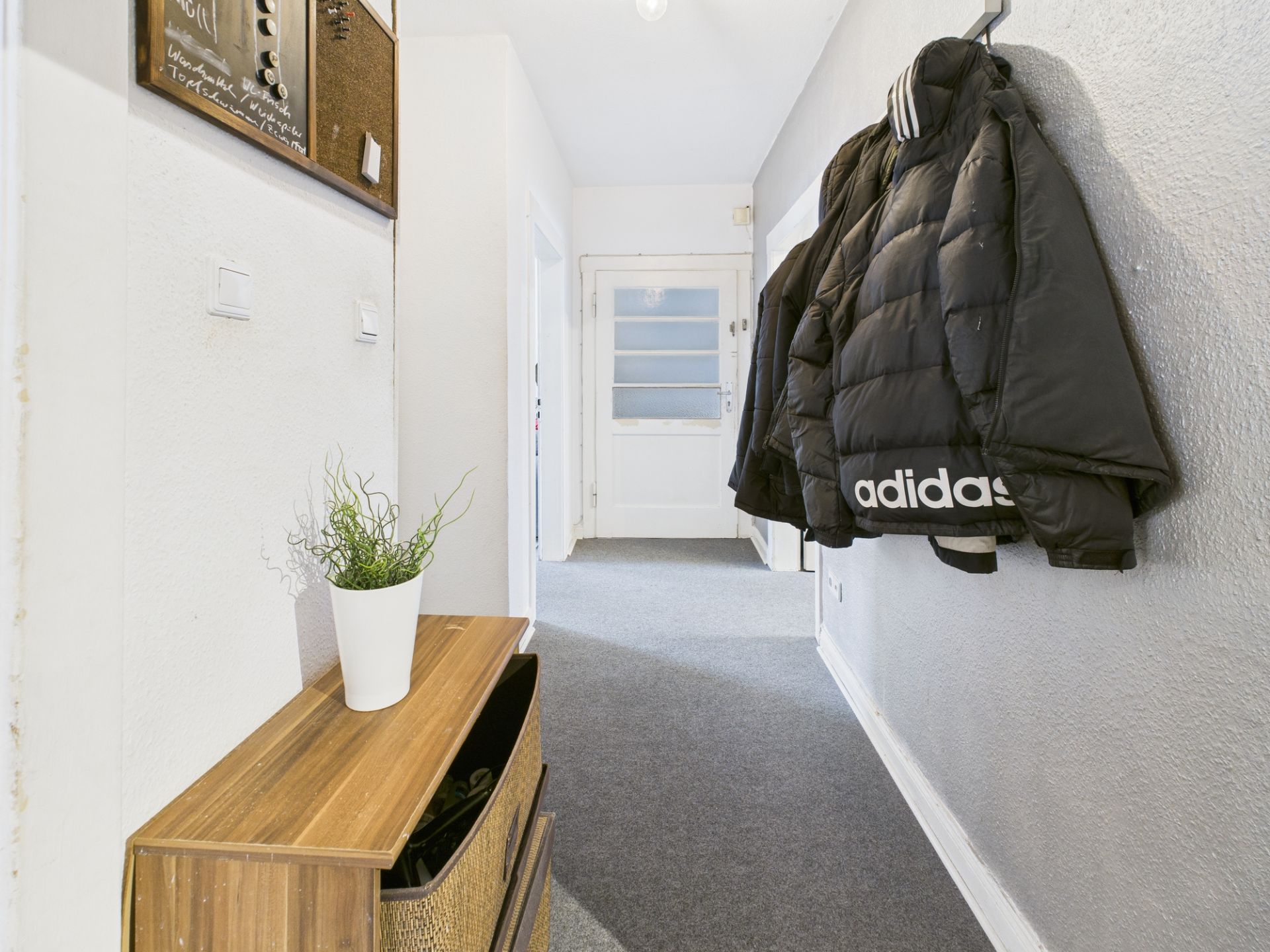 Heller Flur mit Teppichboden, Holzkommode, Pflanze und einer Jacke an der Wand.