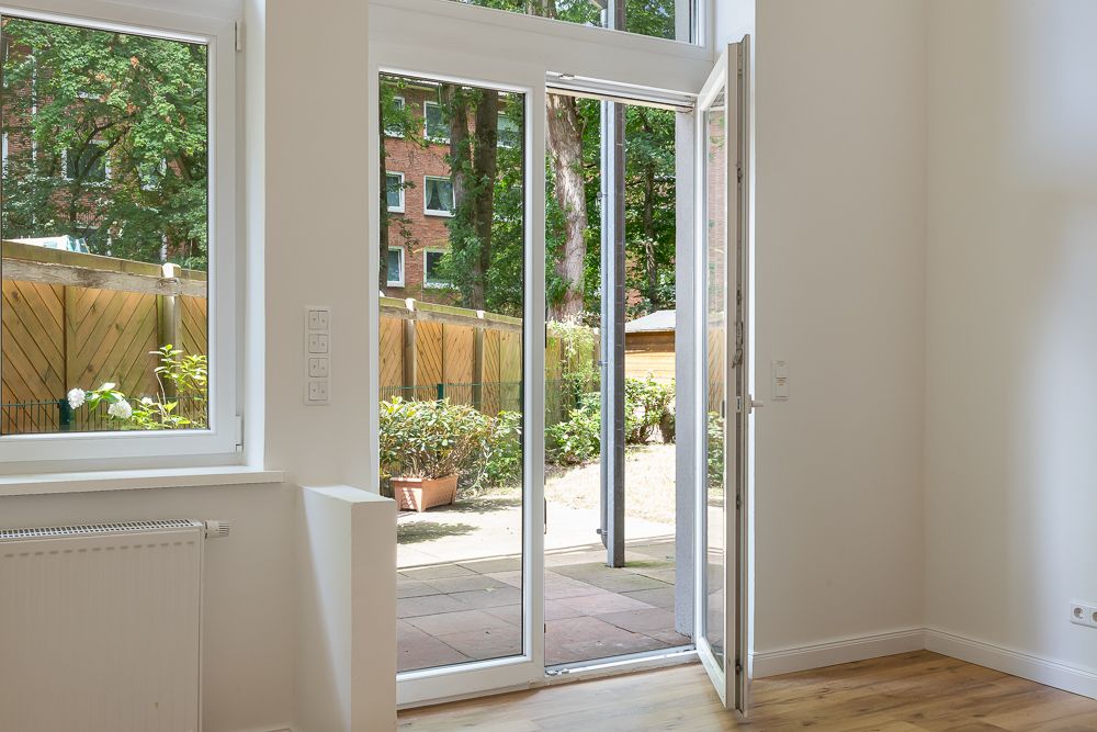 Große Glastür in einem hellen Raum öffnet sich auf eine Terrasse mit Garten und Holzzaun.