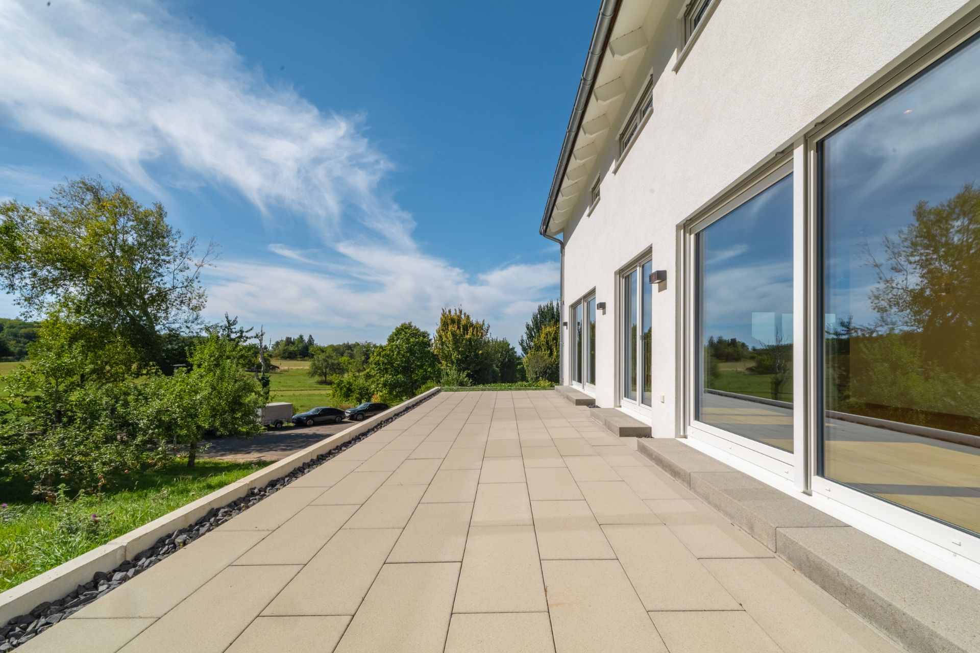 Große Terrasse mit hellen Fliesen neben einem modernen Haus, umgeben von Bäumen und Wiese.