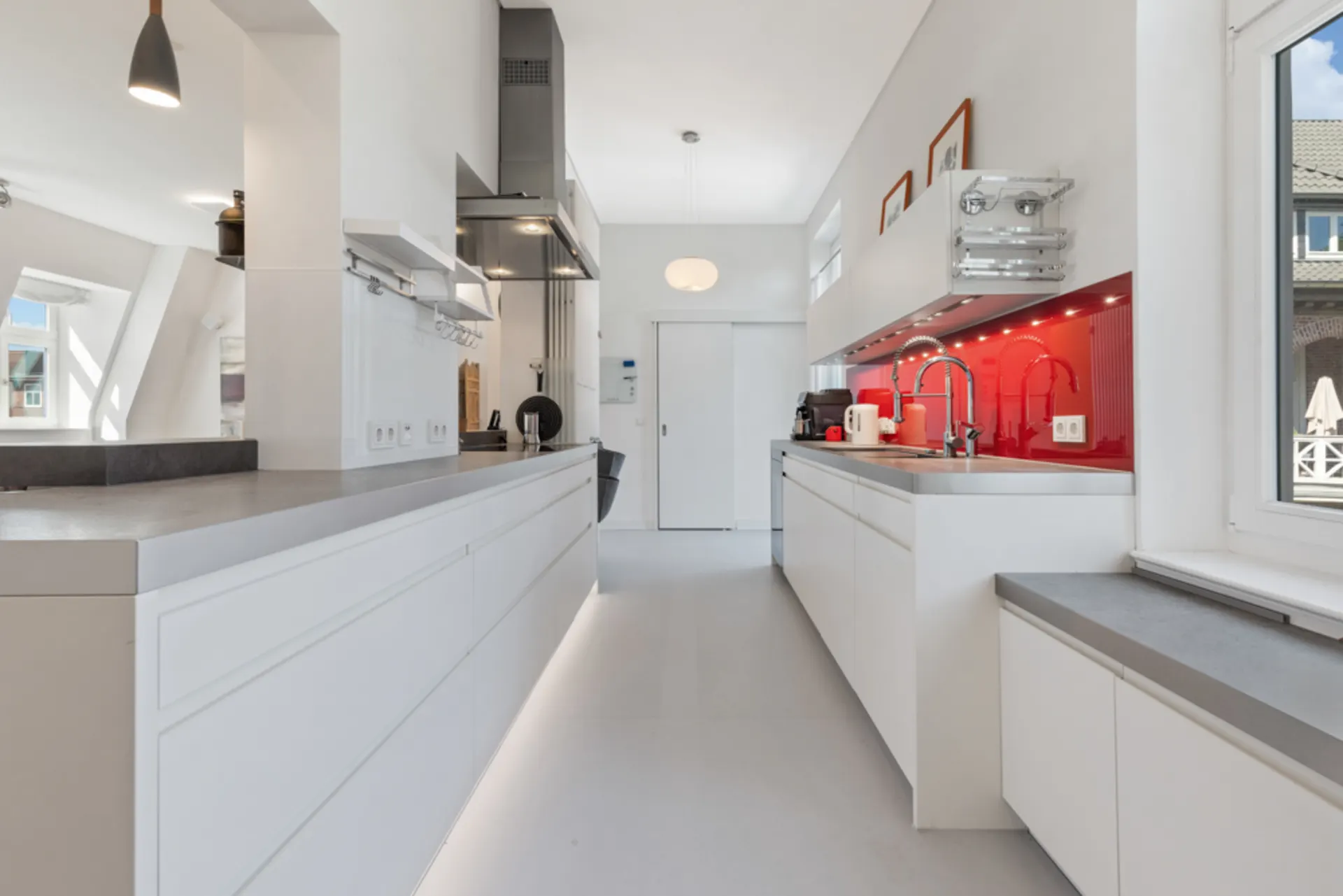 Moderne weiße Küche mit roter Rückwand, großen Fenstern und grauen Arbeitsplatten.