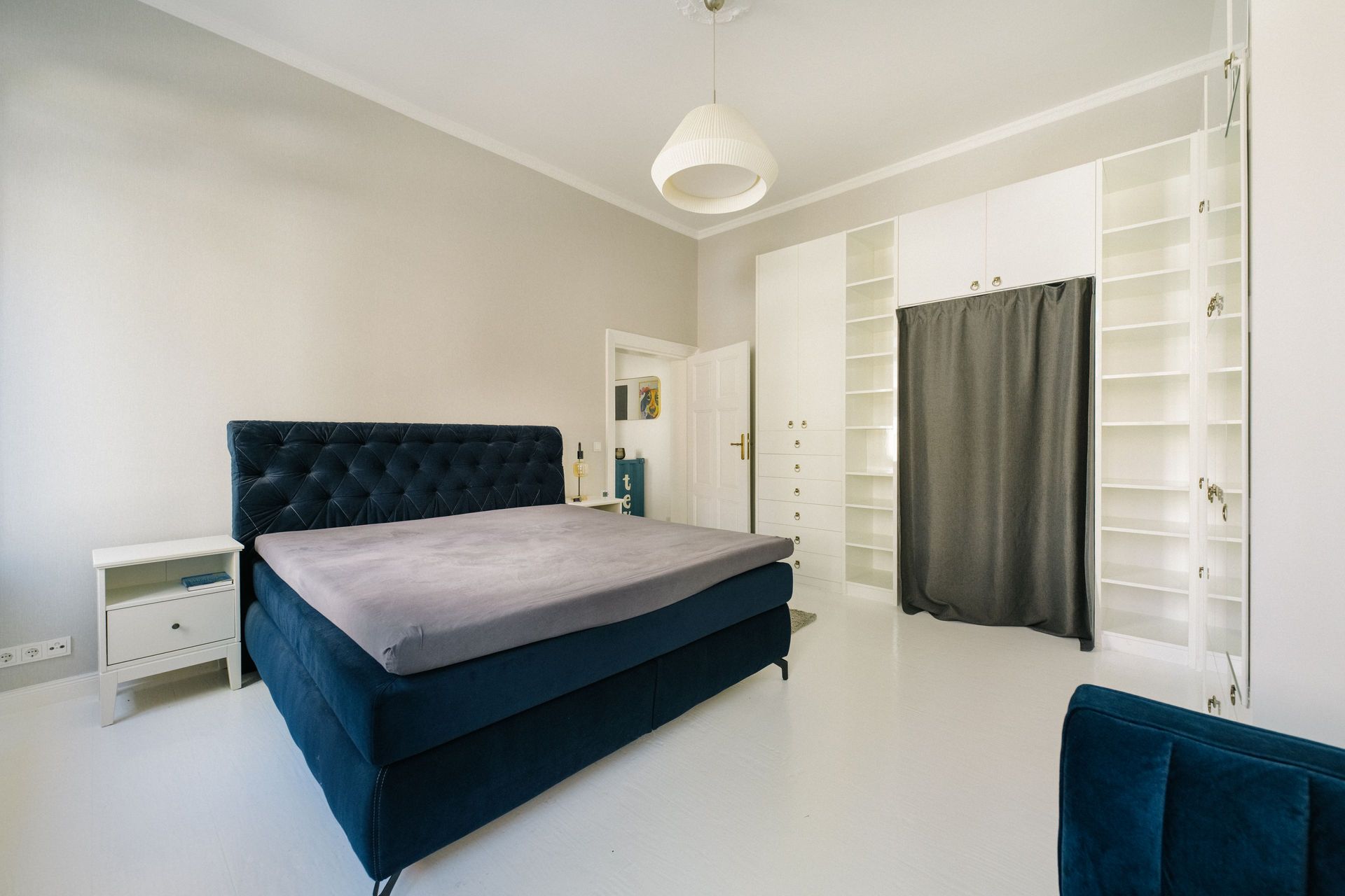 Helles Schlafzimmer mit dunkelblauem Bett, weißen Wänden und Einbauschrank mit Vorhang.