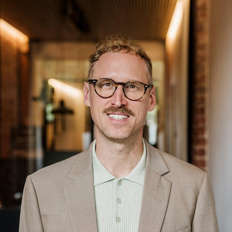 Ein Mann mit Brille und Schnurrbart steht in einem Flur mit Holzwänden.