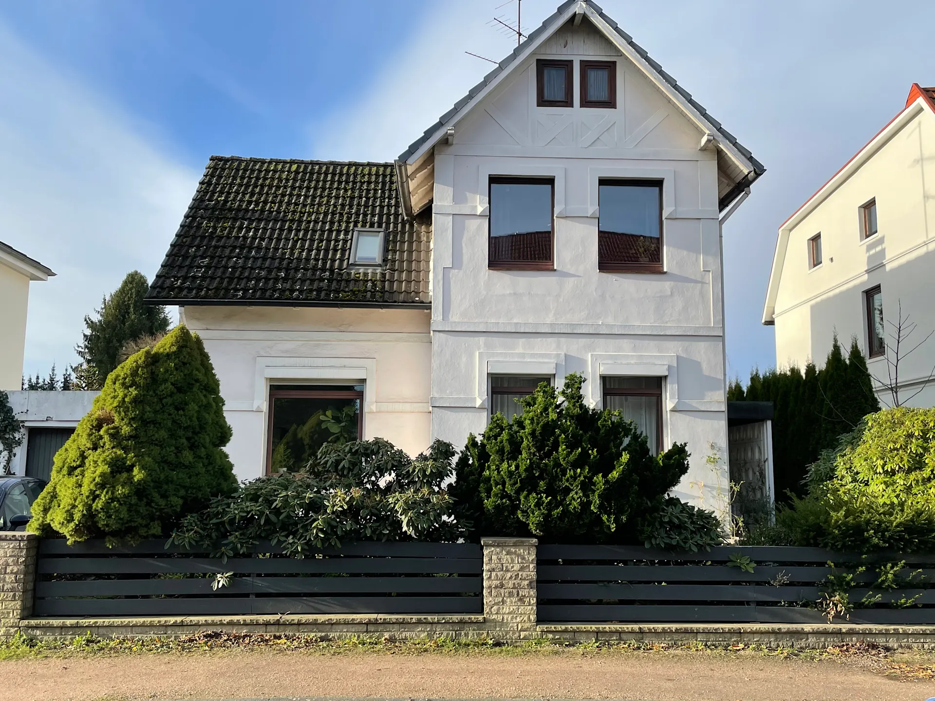 Weißes Wohnhaus mit dunklem Dach, vier Fenstern und vorgelagertem Garten hinter einem Zaun.