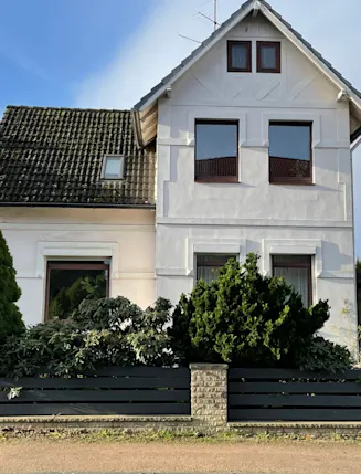 Weißes Wohnhaus mit dunklem Dach, vier Fenstern und vorgelagertem Garten hinter einem Zaun.