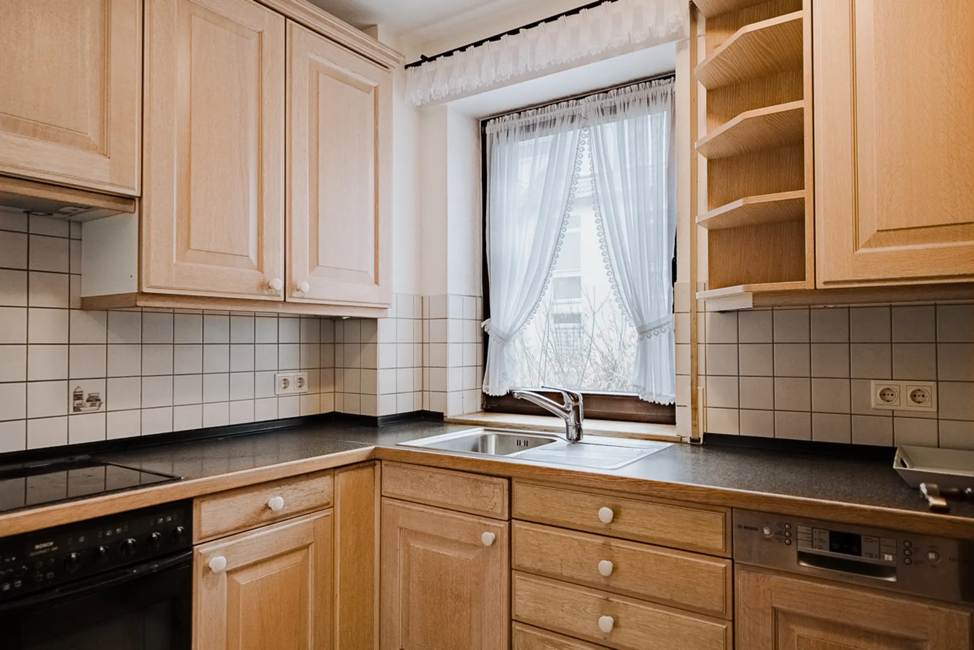 Helle Küche mit Holzschränken, Spüle unter Fenster und weißen Fliesen an der Wand.