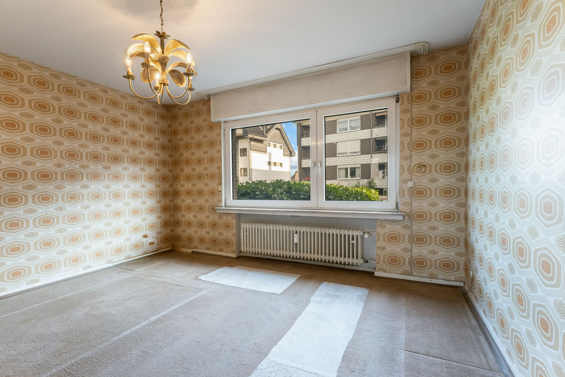 Leeres Zimmer mit gemusterter Tapete, großem Fenster und Kronleuchter.