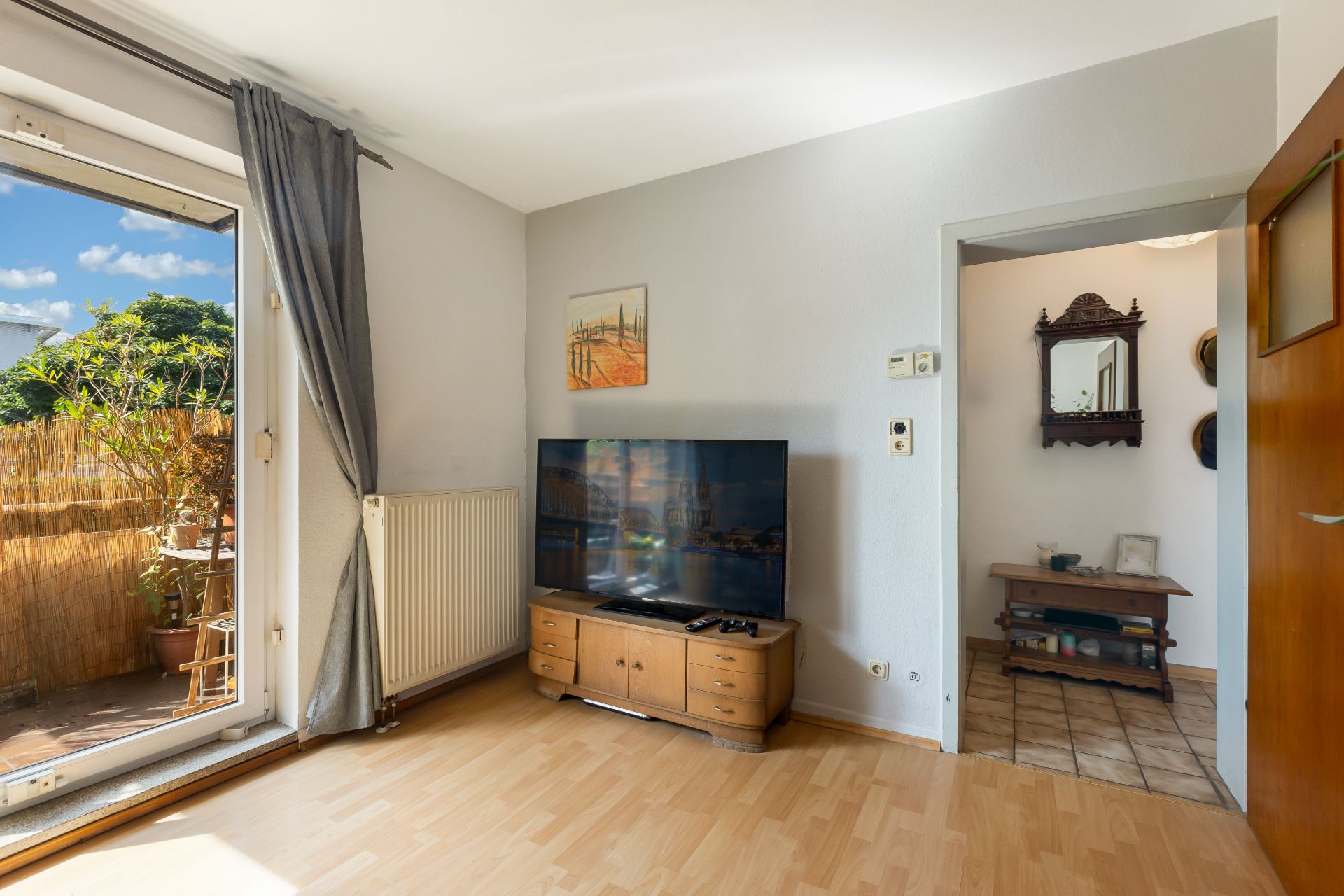 Wohnzimmer mit Fernseher, Holzkommode, offener Tür zum Flur und Zugang zu einem Balkon mit Pflanzen.