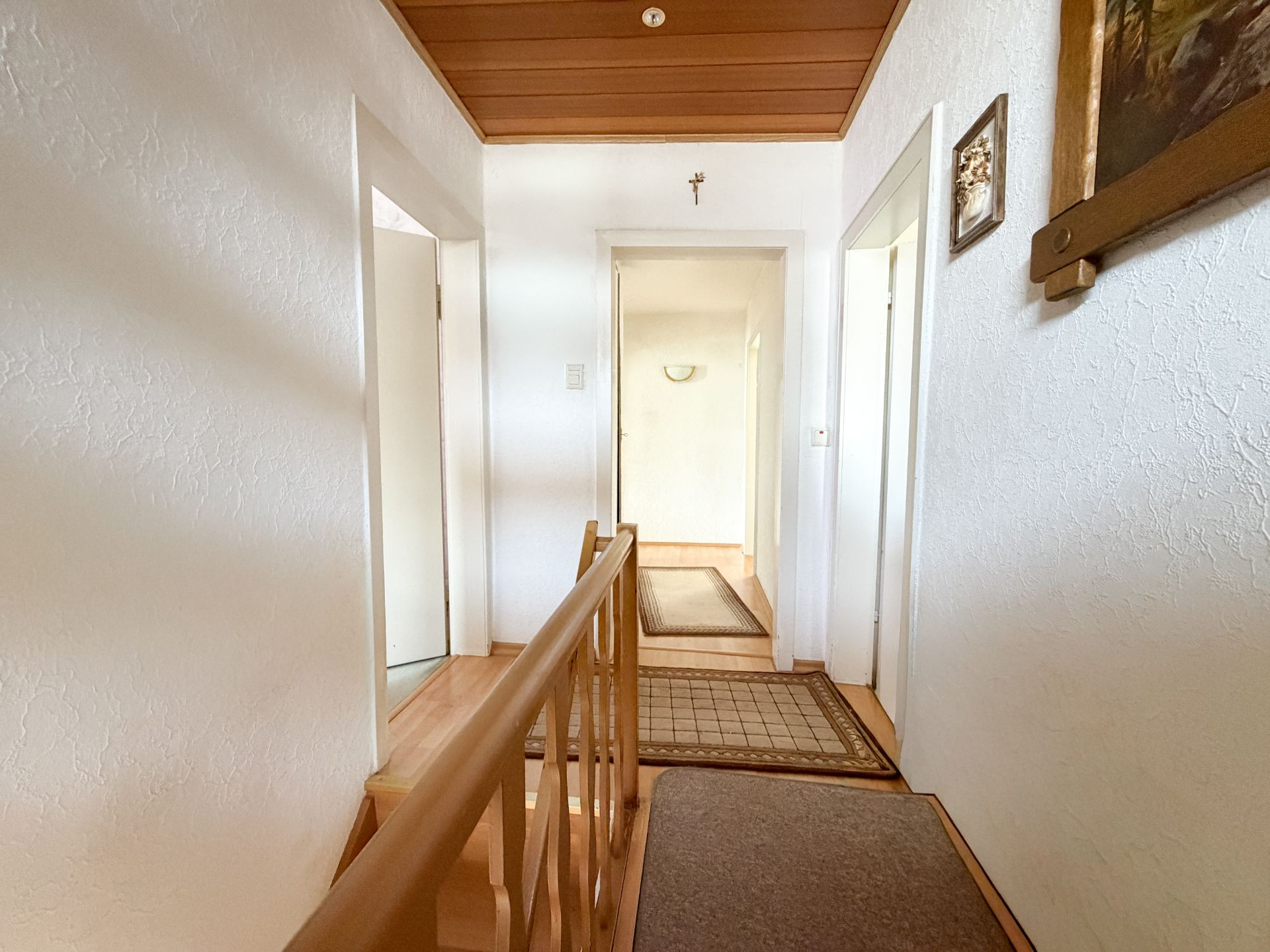 Heller Flur mit Holzgeländer, mehreren Türen und Teppichen, weiße Wände, Holzdecke.