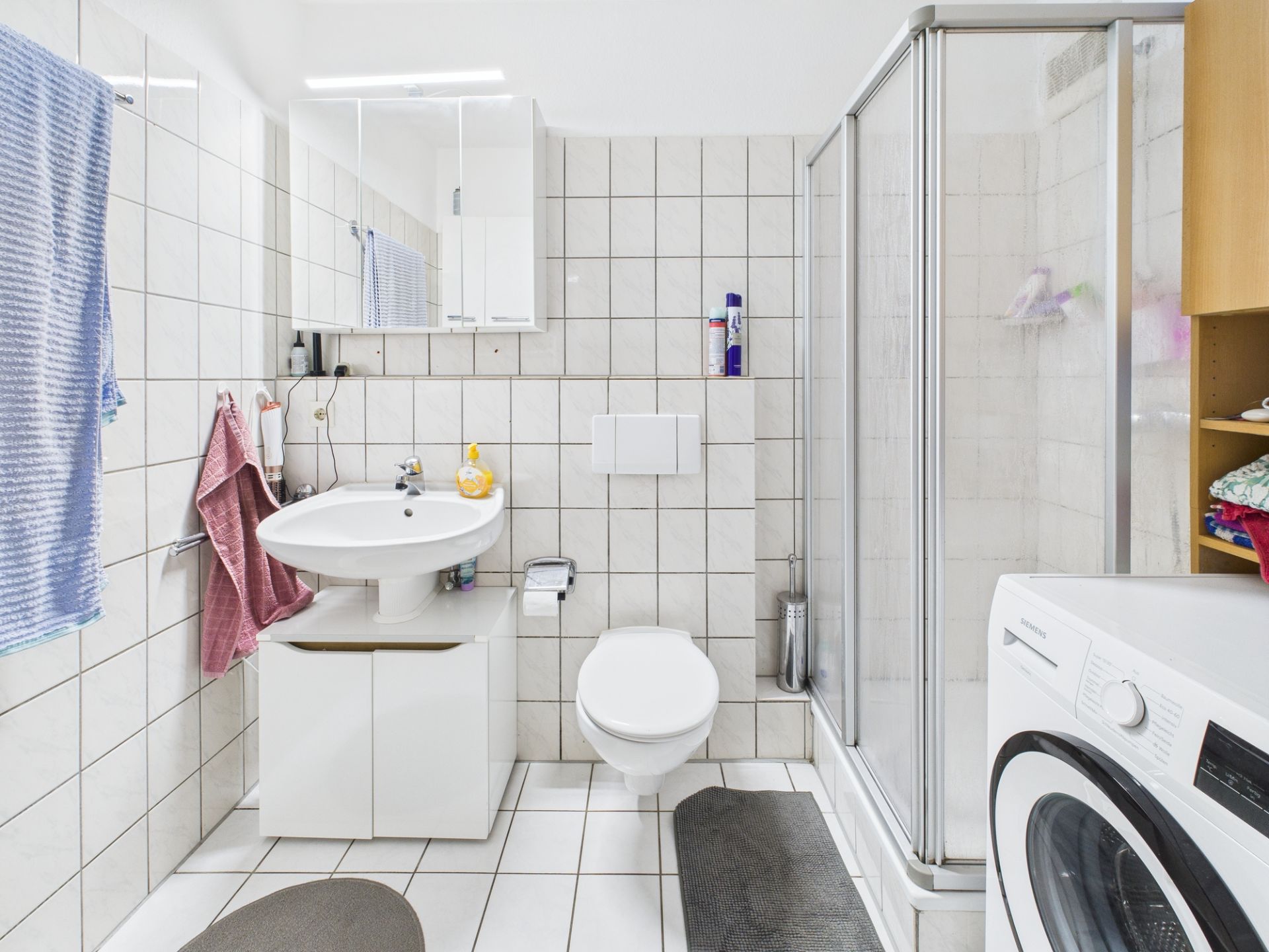 Modernes Badezimmer mit Dusche, Waschmaschine, Waschbecken, WC und Fliesenboden.
