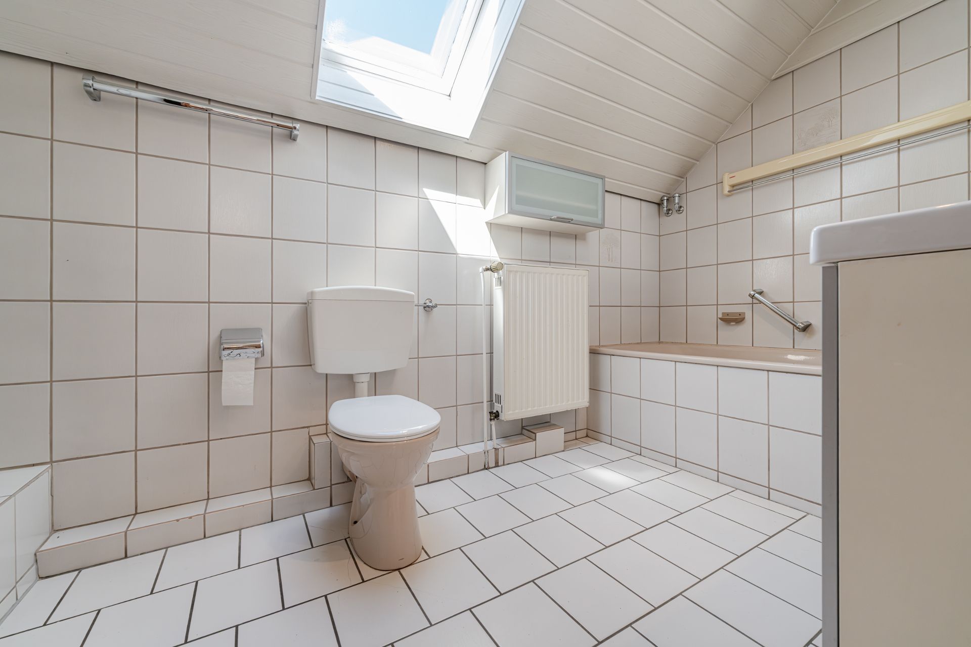 Helles Badezimmer mit WC, Badewanne und Dachfenster, gefliest in Weiß.