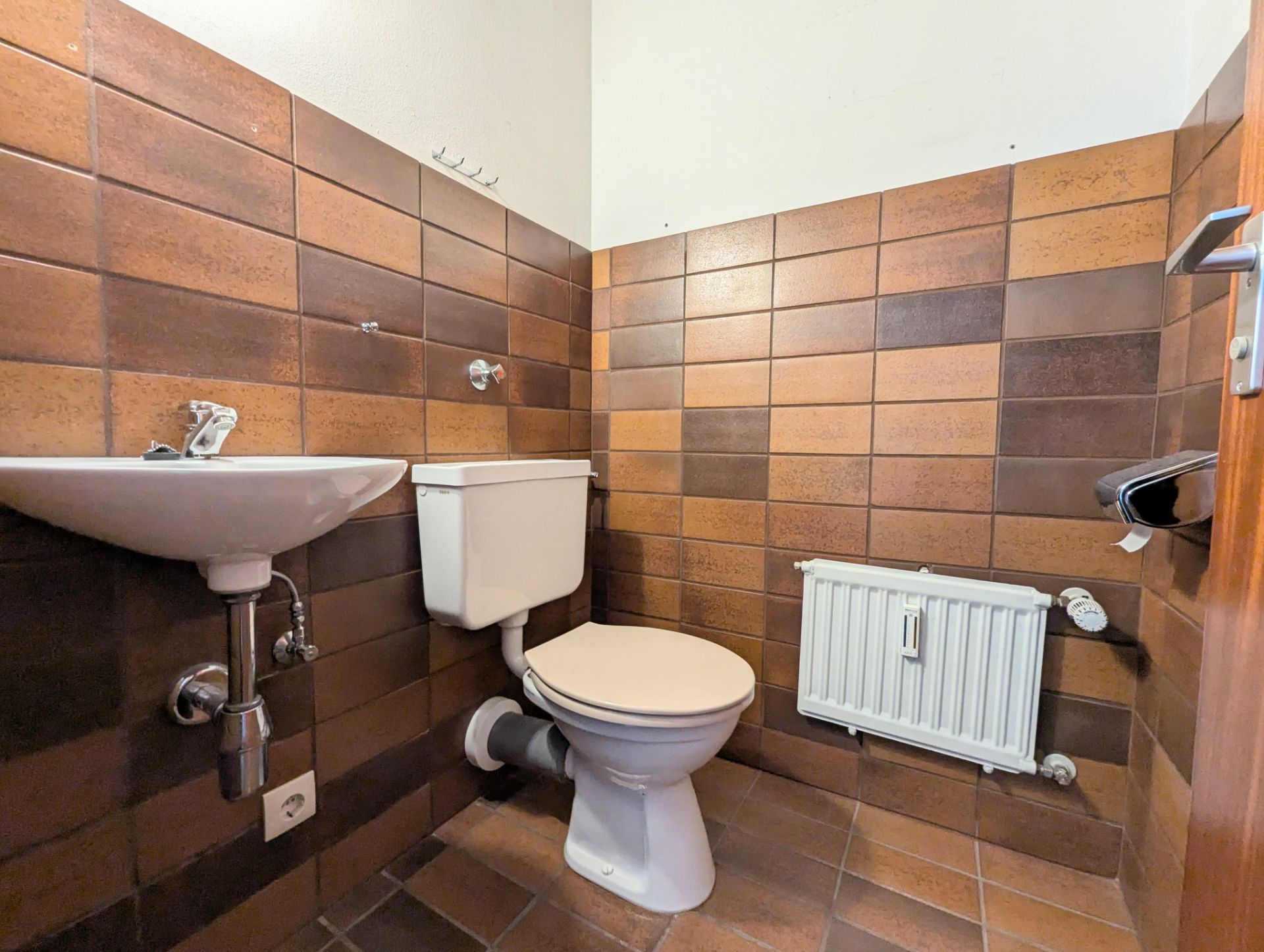 Kleines Bad mit braunen Fliesen, WC, Waschbecken und Heizkörper.