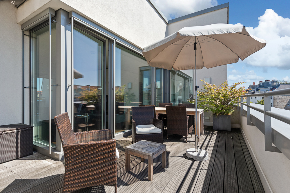 Große Terrasse mit Sitzmöbeln, Tisch und Sonnenschirm, umgeben von Glas und Pflanzen.
