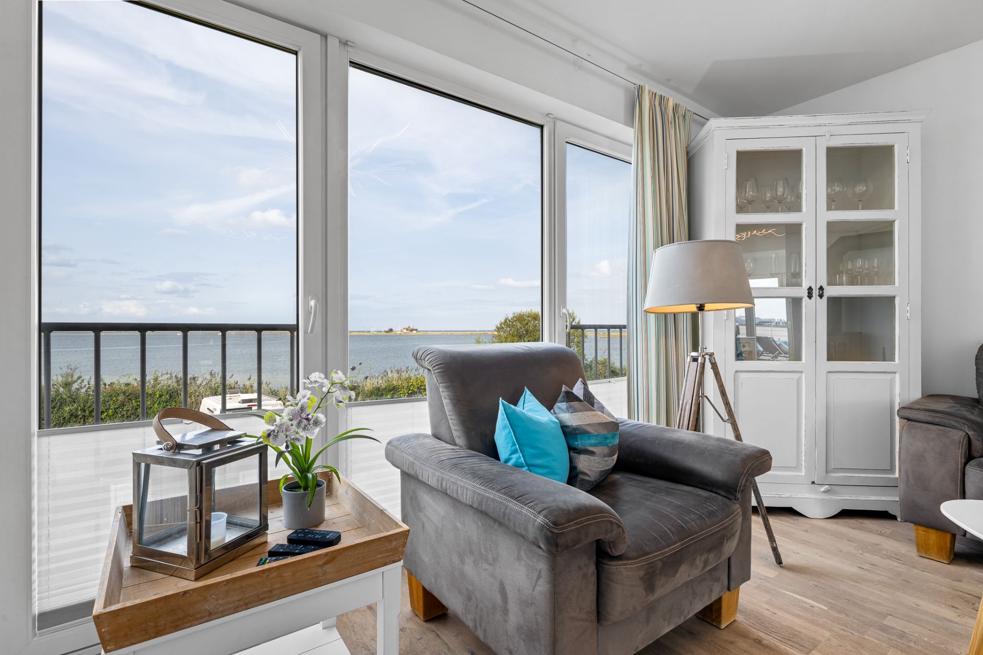 Gemütliches Wohnzimmer mit Sessel, Beistelltisch, Stehlampe und Blick aufs Wasser durch große Fenster.