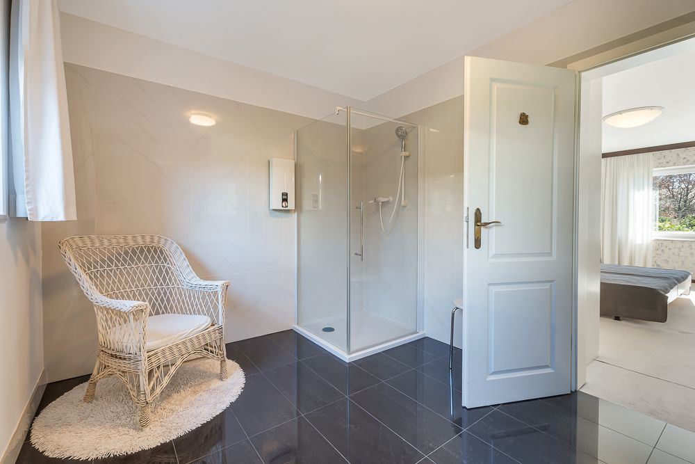 Modernes Badezimmer mit Glasdusche, weißem Korbstuhl und glänzendem dunklen Fliesenboden.