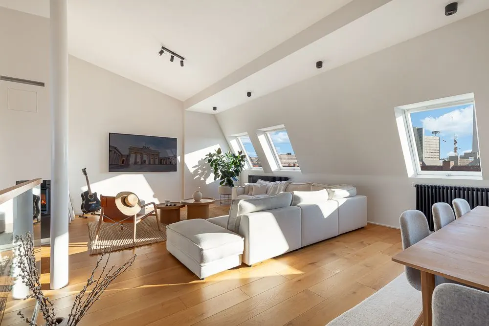Helles Wohnzimmer mit weißem Sofa, Holzboden, Fernseher, Gitarre und großen Fenstern.