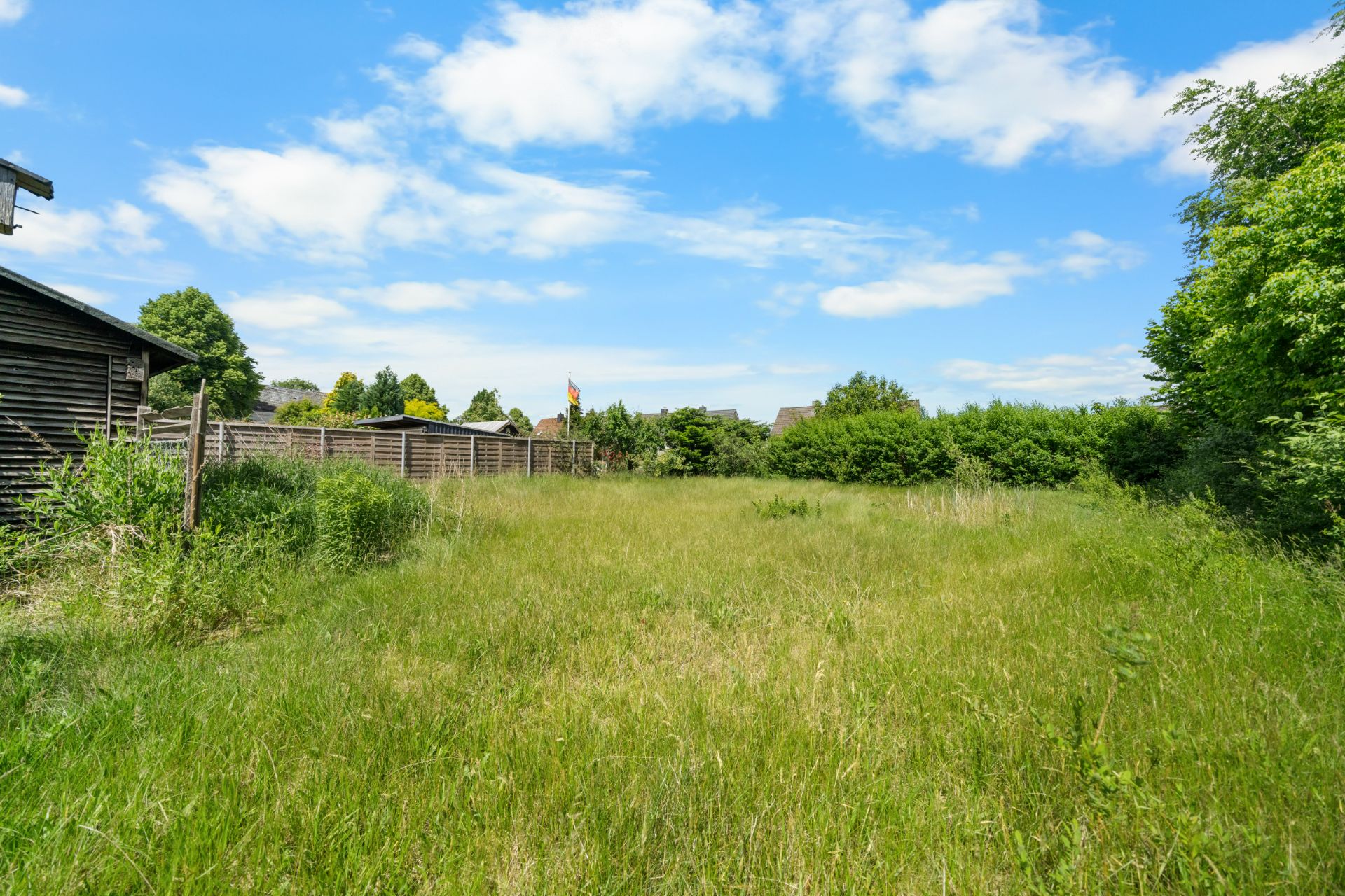 Grünes Grundstück mit Wiese, Holzzaun und Büschen an den Seiten unter blauem Himmel.