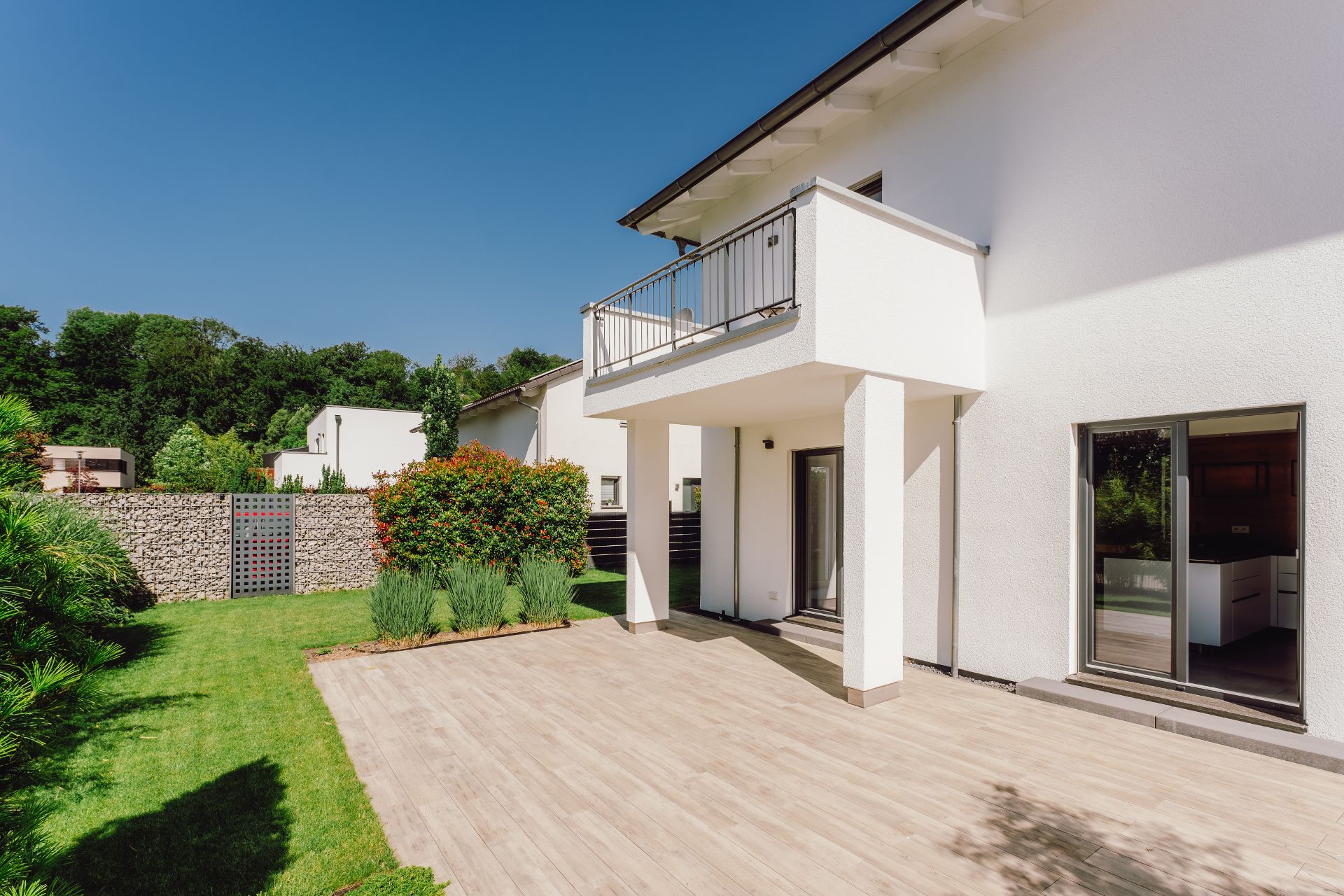 Moderne Terrasse mit Garten und Balkon an einem weißen Wohnhaus bei Sonnenschein.