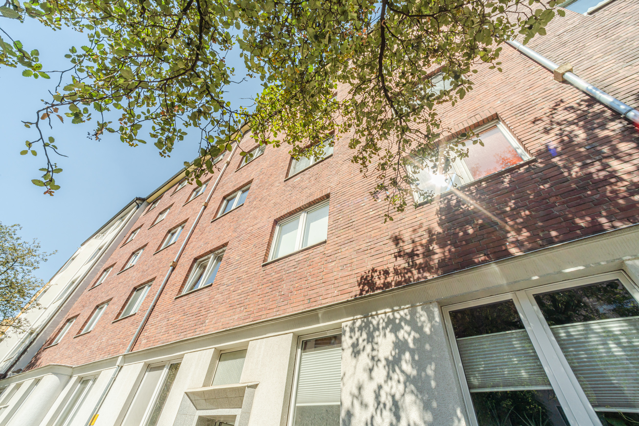 Mehrstöckiges Wohnhaus mit roter Klinkerfassade und vielen Fenstern, Äste und Blätter im Vordergrund.