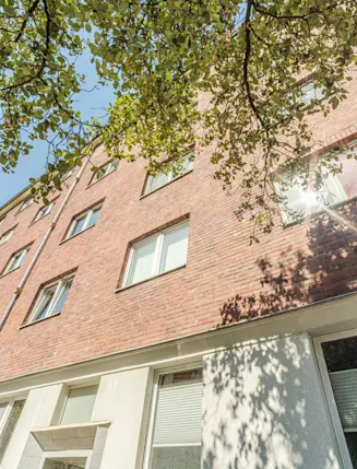 Mehrstöckiges Wohnhaus mit roter Klinkerfassade und vielen Fenstern, Äste und Blätter im Vordergrund.